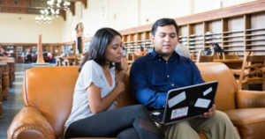 Two students look at a laptop together in a library.