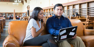 Two students look at a laptop together in a library.