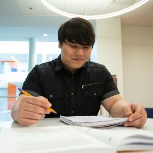 A student smiles while looking down at a notebook and holding a pencil