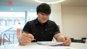 A student smiles while looking down at a notebook and holding a pencil
